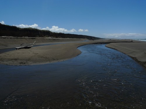 A winding river flows through a sandy beach under a clear blue sky, with distant cliffs in the background.