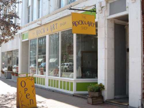 A storefront with a yellow sign reading "The Rock & Art Shop" and an open sign outside, featuring large windows.