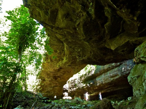 Two people stand beneath a large rock overhang in a lush, green forest setting. Sunlight filters through the trees.