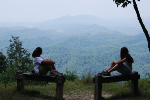 Two girls sit on benches, gazing at a misty mountain landscape with lush green hills in the background.