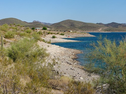 A serene lakeside view with rocky shorelines, green shrubs, and distant hills under a clear blue sky.