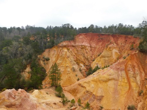 A colorful canyon with layered orange and yellow rock formations, surrounded by green trees under a cloudy sky.