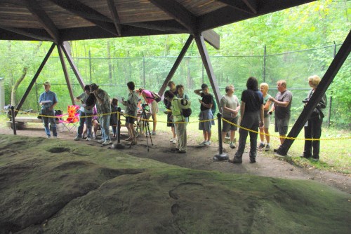 A group of people observing and photographing a rock formation under a shelter in a wooded area.