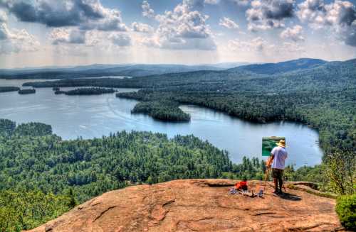 A painter stands on a rocky overlook, capturing a scenic view of a lake surrounded by lush green forests and mountains.