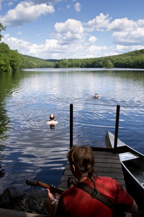 A person plays guitar on a dock while two people swim in a calm lake surrounded by lush greenery and blue skies.