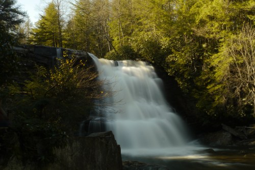 A serene waterfall cascades over rocks, surrounded by lush green trees in a tranquil forest setting.