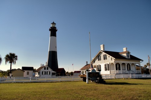 A tall lighthouse beside a white house, surrounded by green grass and a clear blue sky.