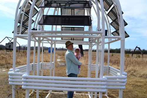 A man stands inside a white structure holding a child, surrounded by a grassy field under a clear sky.