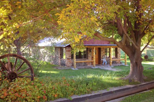 A charming stone cottage surrounded by greenery and trees, with a vintage wagon wheel in the foreground.