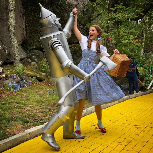 A woman in a blue dress dances with a silver tin man on a yellow brick road surrounded by greenery.