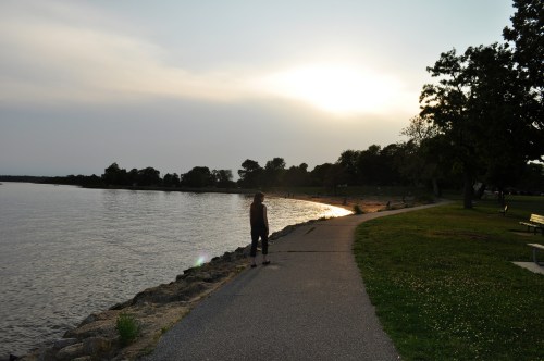 A person stands by a calm waterfront at sunset, with trees and a path along the shore.