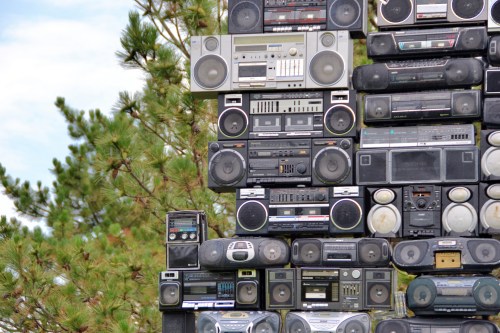 A towering stack of vintage boomboxes against a backdrop of trees and blue sky.