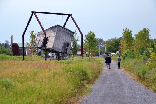 Two people walk along a path beside a tilted wooden structure supported by a metal frame, surrounded by greenery.