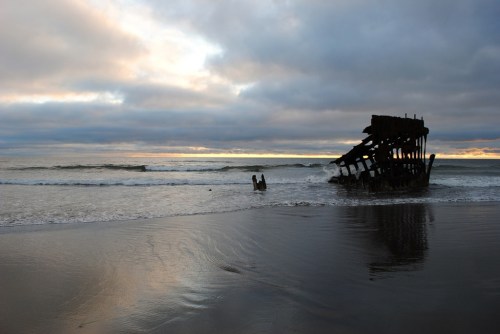 A shipwreck partially submerged in calm waters at sunset, with cloudy skies reflecting on the wet sand.