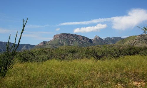 A scenic view of green hills and a prominent rocky mountain under a clear blue sky with scattered clouds.