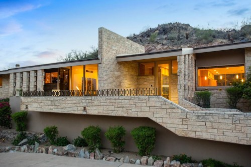 Modern stone house with large windows, set against a rocky hillside and landscaped greenery.