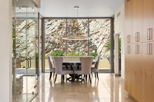 Modern dining area with a round table and gray chairs, featuring a rock wall view through large glass windows.