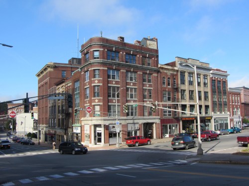 Historic brick buildings line a street, with cars parked and traffic lights visible under a clear blue sky.