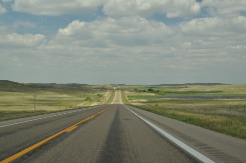 A long, straight road stretches into the distance, surrounded by green fields and a blue sky with fluffy clouds.