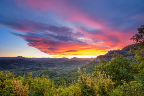 Vibrant sunset over a mountainous landscape, with colorful clouds and lush greenery in the foreground.