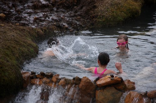 Three children play in a natural hot spring, splashing water and enjoying the warm surroundings.