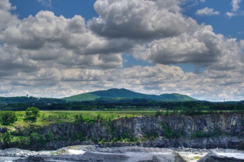 A scenic view of rolling green hills under a cloudy sky, with a rocky foreground and a river in the distance.