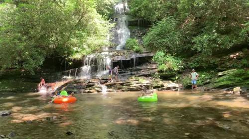 A serene waterfall cascades into a pool, with people enjoying the water on colorful inner tubes surrounded by lush greenery.