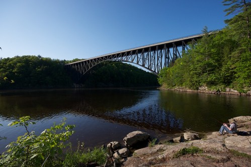 A large arch bridge spans a calm river, surrounded by lush green trees and rocky banks under a clear blue sky.