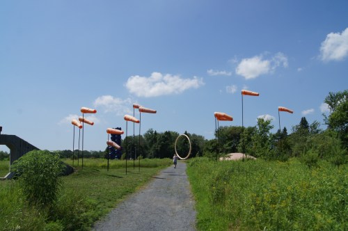 A pathway through a grassy area with colorful sculptures and flags against a blue sky.