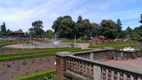 A scenic park with a fountain, lush greenery, and flower beds under a clear blue sky.