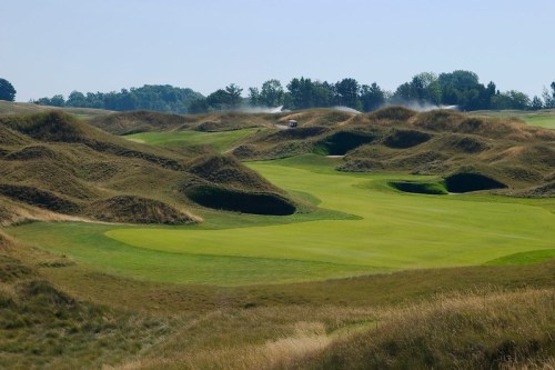 A scenic golf course with rolling hills, lush green grass, and a clear blue sky in the background.