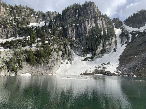 A serene mountain lake surrounded by rocky cliffs and patches of snow, reflecting the landscape in calm waters.
