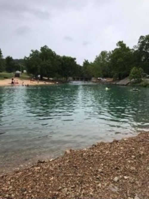 A serene lake surrounded by trees, with a sandy beach area in the background under a cloudy sky.