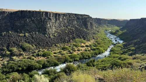 A winding river flows through a lush green valley surrounded by steep, rocky cliffs under a clear blue sky.