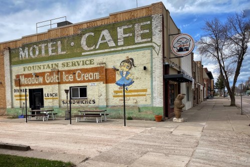 A vintage motel and café building with a faded mural, featuring outdoor seating and a bear statue on the sidewalk.