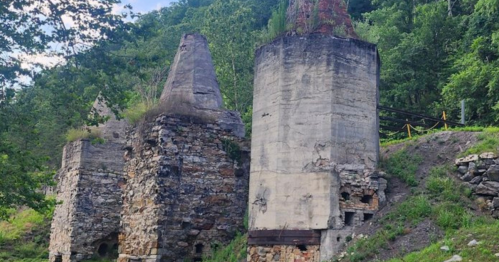 Ruins of old stone structures surrounded by greenery, with trees and a hillside in the background.