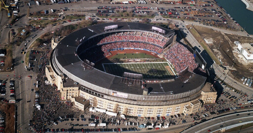 Aerial view of a large stadium filled with fans, surrounded by parking lots and a river in the background.