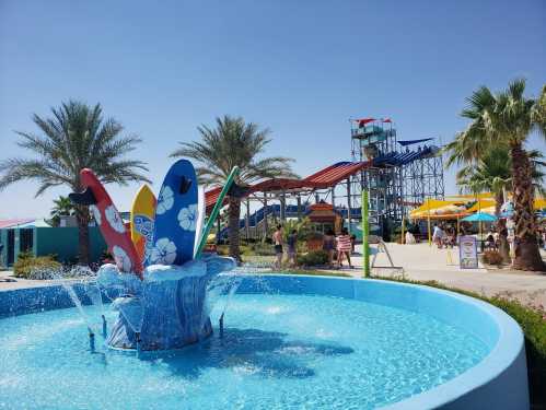 A colorful water park scene featuring a fountain with surfboards and a tall water slide in the background, surrounded by palm trees.