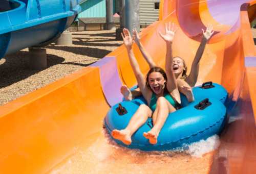 Two girls joyfully slide down a colorful water slide on a blue inner tube, splashing into the water below.