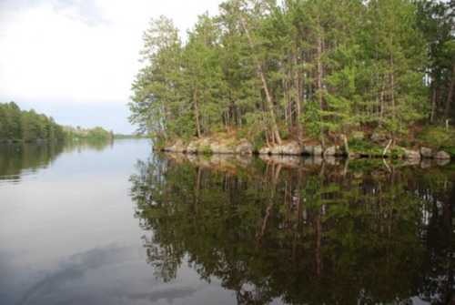 A serene lake scene with trees reflecting on the calm water, surrounded by rocky shores and a cloudy sky.