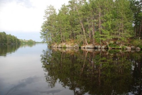 A serene lake scene with trees reflecting on the calm water, surrounded by rocky shores and a cloudy sky.
