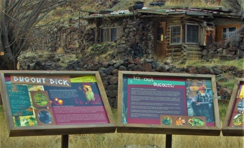 A rustic cabin surrounded by rocks, with informational signs about Dugout Dick and ice caves in the foreground.