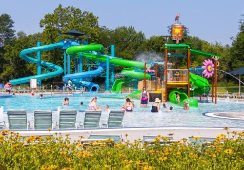 A vibrant water park with slides, a splash pad, and families enjoying the pool on a sunny day surrounded by flowers.