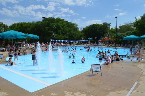 A busy outdoor pool with people swimming and playing, surrounded by lounge chairs and umbrellas under a blue sky.