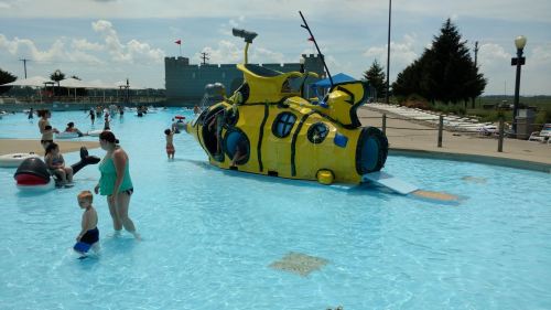 A colorful yellow submarine play structure in a pool with children and adults enjoying the water on a sunny day.