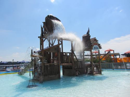 A water play structure with a large bucket pouring water into a pool, surrounded by slides and sunny skies.