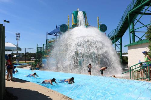 Children play in a water park, sliding down a blue slide with a large water feature splashing in the background.