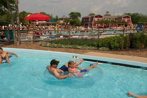 A man and a child float on a tube in a pool, with a busy water park in the background and people enjoying the sun.