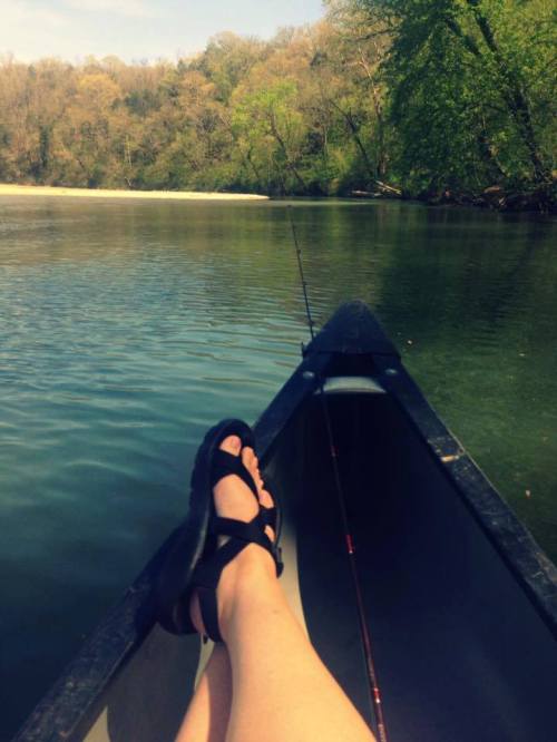 A person relaxes in a kayak, feet propped up, with a fishing rod beside them and a serene river landscape in the background.