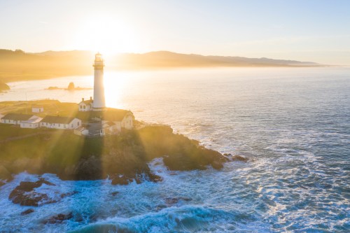 A lighthouse stands on a rocky shore, illuminated by the sun, with waves crashing against the coast at sunset.